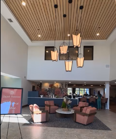 Bright senior living lobby with a high wood-paneled ceiling, hanging pendant lights, a seating area of pink armchairs around a round table, and a 'Saint Therese' welcome sign.