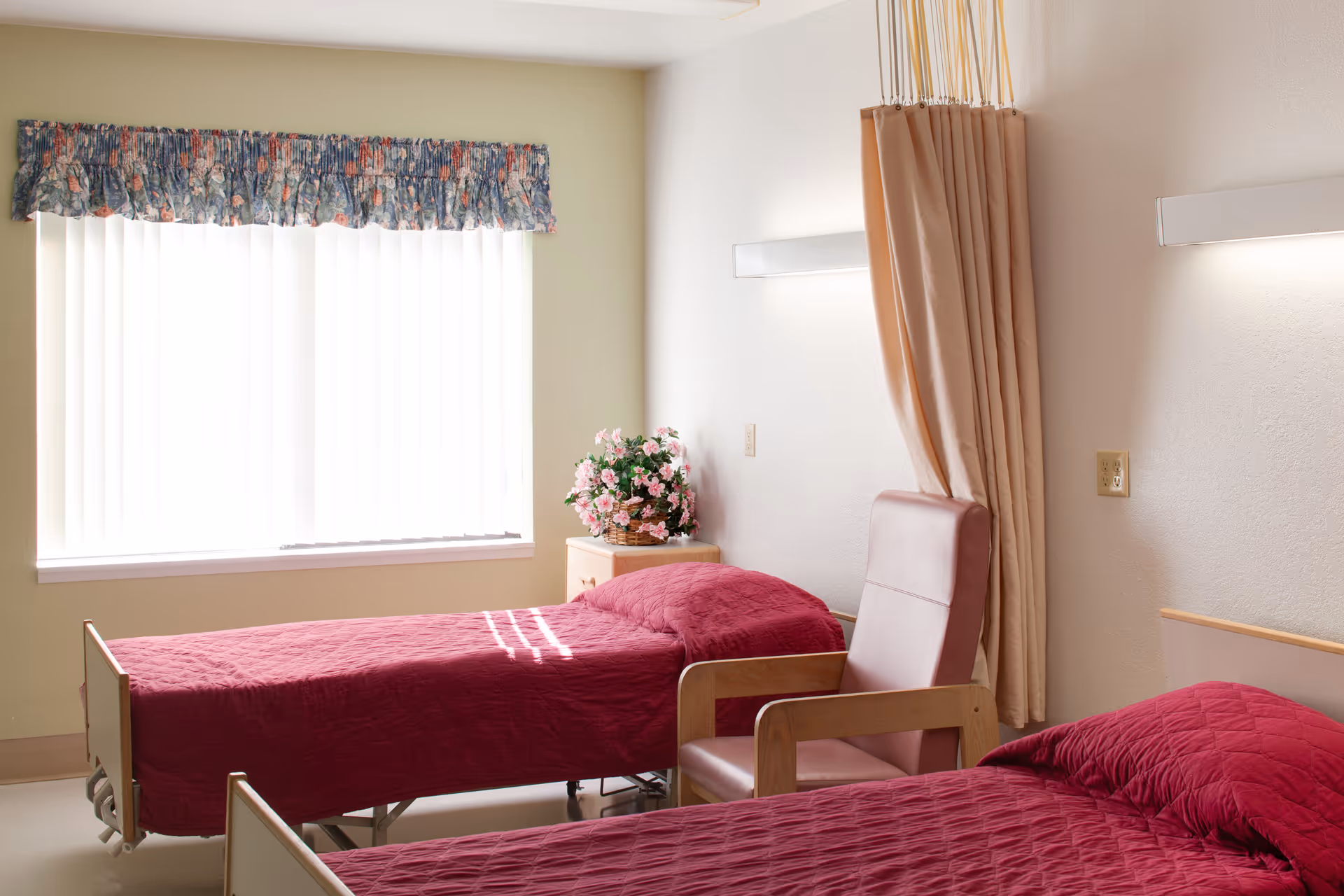 A bright room with two single beds covered in red quilts, a padded chair between them, a small wooden nightstand with a basket of pink flowers, vertical blinds on the window with a floral valance, and a beige privacy curtain partially drawn.