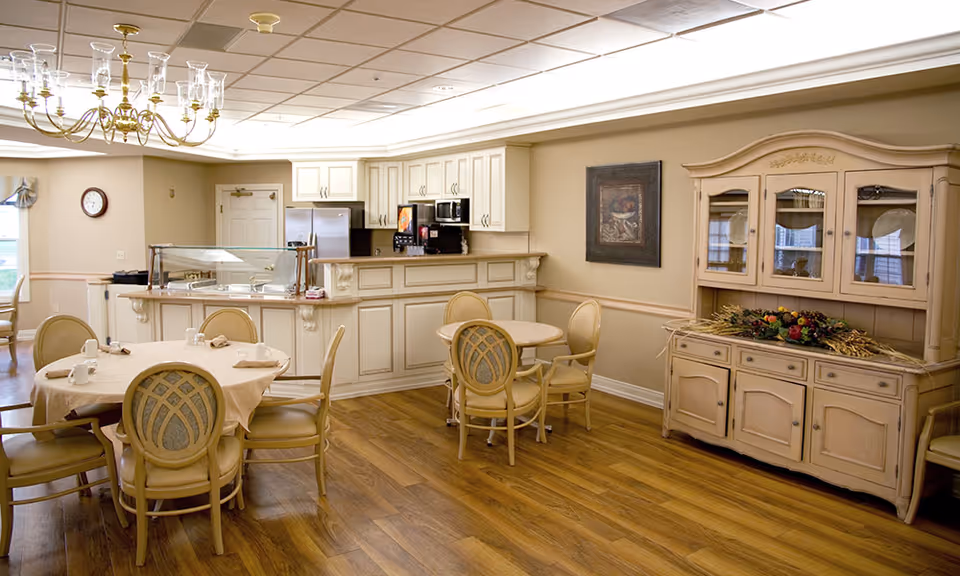 A cozy dining area in a senior living facility featuring round tables with beige tablecloths and chairs, a wooden hutch decorated with a floral arrangement, a kitchen counter with a beverage dispenser, and a chandelier overhead. The room has warm lighting and wooden flooring.