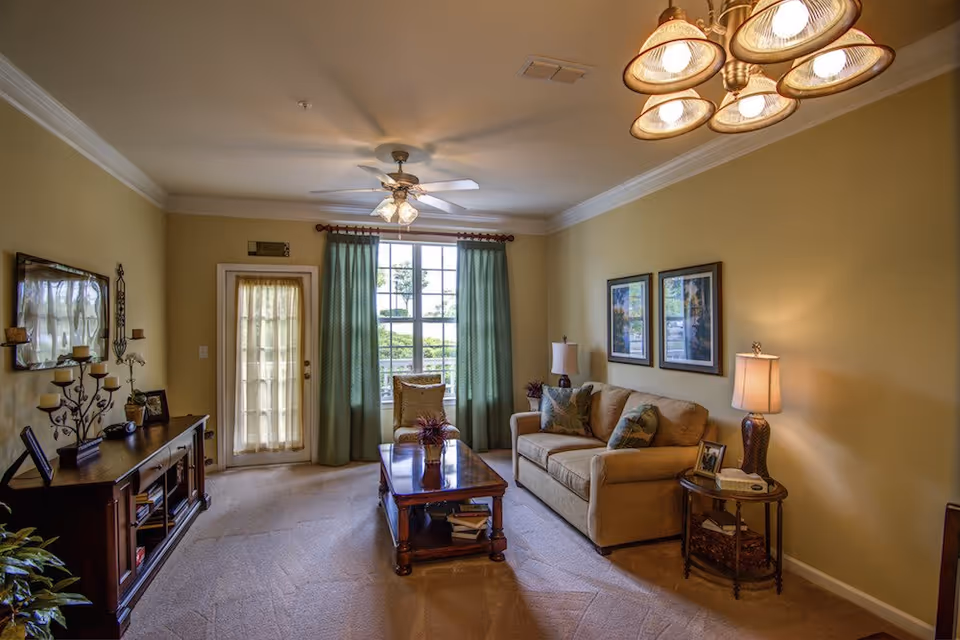 A cozy living room with beige walls and carpet, featuring a beige sofa with patterned pillows, a wooden coffee table with decorative plants and books, a side table with a lamp and framed photo, a wooden media console with candles and decor, a ceiling fan with lights, and a large window with green curtains letting in natural light.