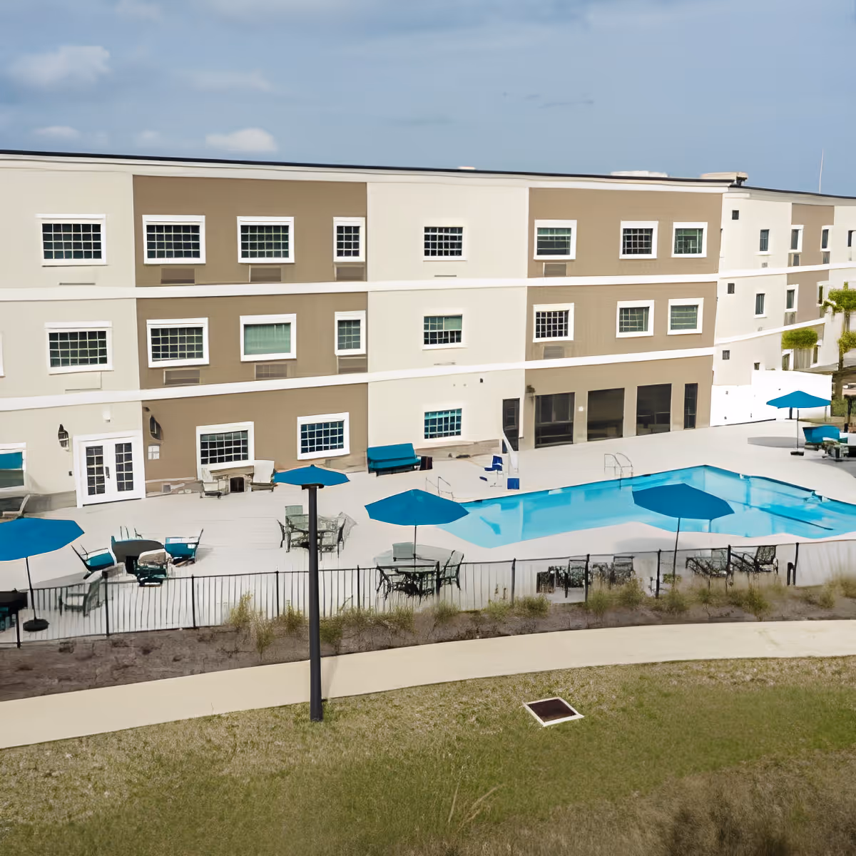 Outdoor view of a multi-story assisted living facility building with a swimming pool surrounded by tables, chairs, and blue umbrellas. There is a paved walkway and some landscaping with grass and plants in the foreground under a partly cloudy sky.