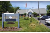 Exterior view of Apple Rehab Colchester facility with a sign in the foreground displaying the facility name. The building has a peaked roof and an American flag is visible near the entrance. There are parked cars and a well-maintained lawn with shrubs and trees.
