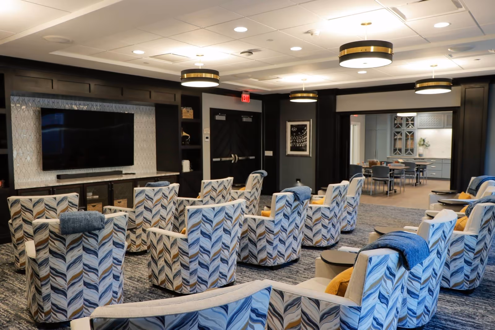 A senior living facility common area with multiple patterned armchairs arranged in rows facing a large flat-screen TV mounted on a dark wood-paneled wall. The room has modern circular ceiling lights, a carpeted floor, and an adjacent dining area visible through a wide doorway.