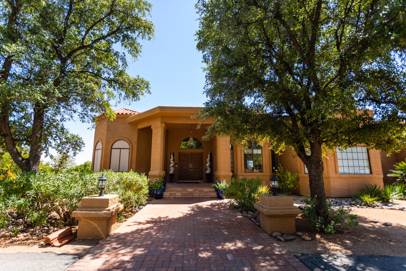 Front exterior view of a single-story building with a tiled roof, large wooden double doors, and columns at the entrance. The pathway leading to the entrance is paved with bricks and flanked by greenery and trees providing shade.