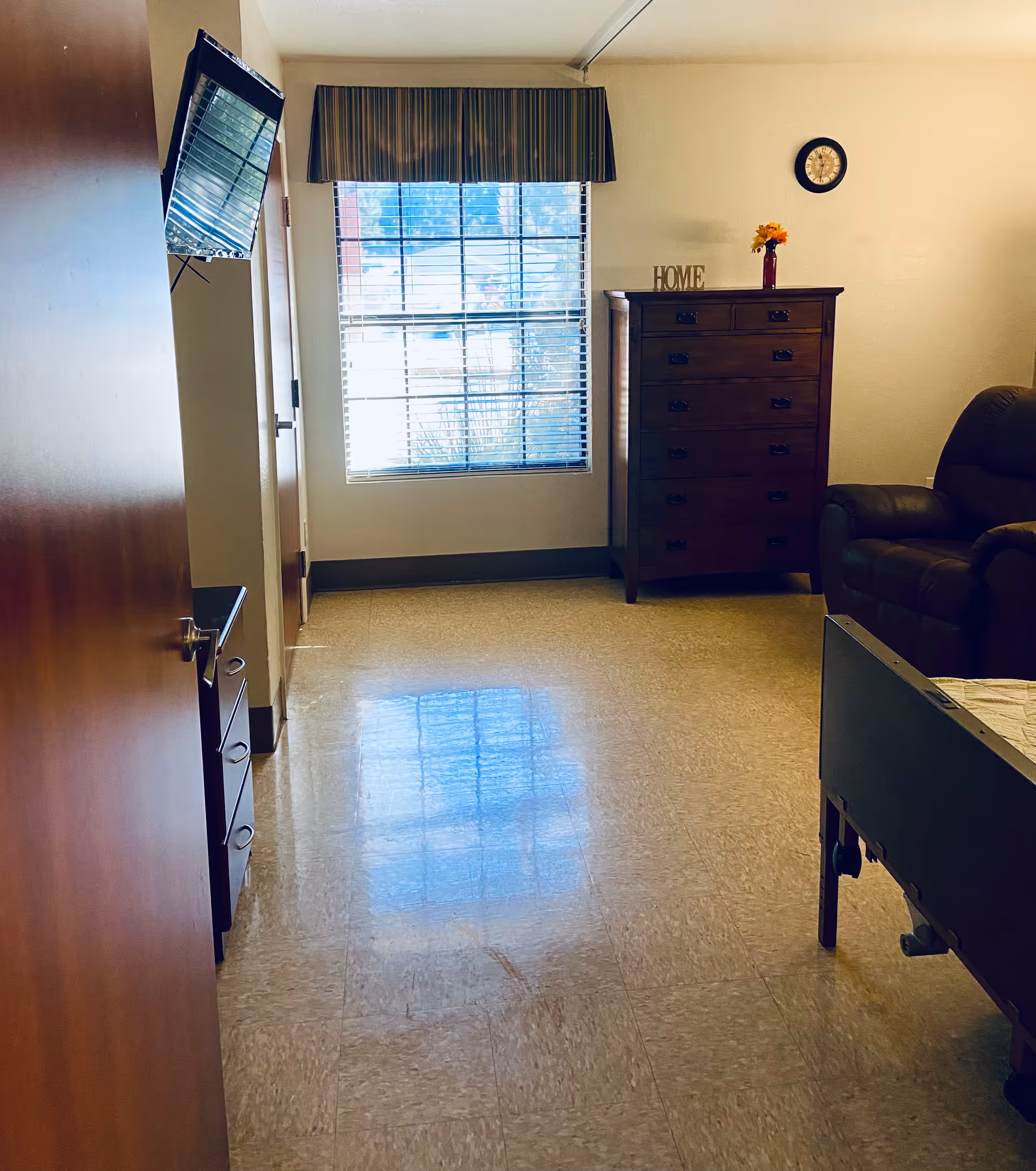A senior living facility room with a window covered by blinds and a valance, a wooden dresser with a vase of flowers and a decorative 'HOME' sign on top, a wall clock, a brown recliner chair, and part of a hospital-style bed visible. The floor is tiled and the walls are light-colored.