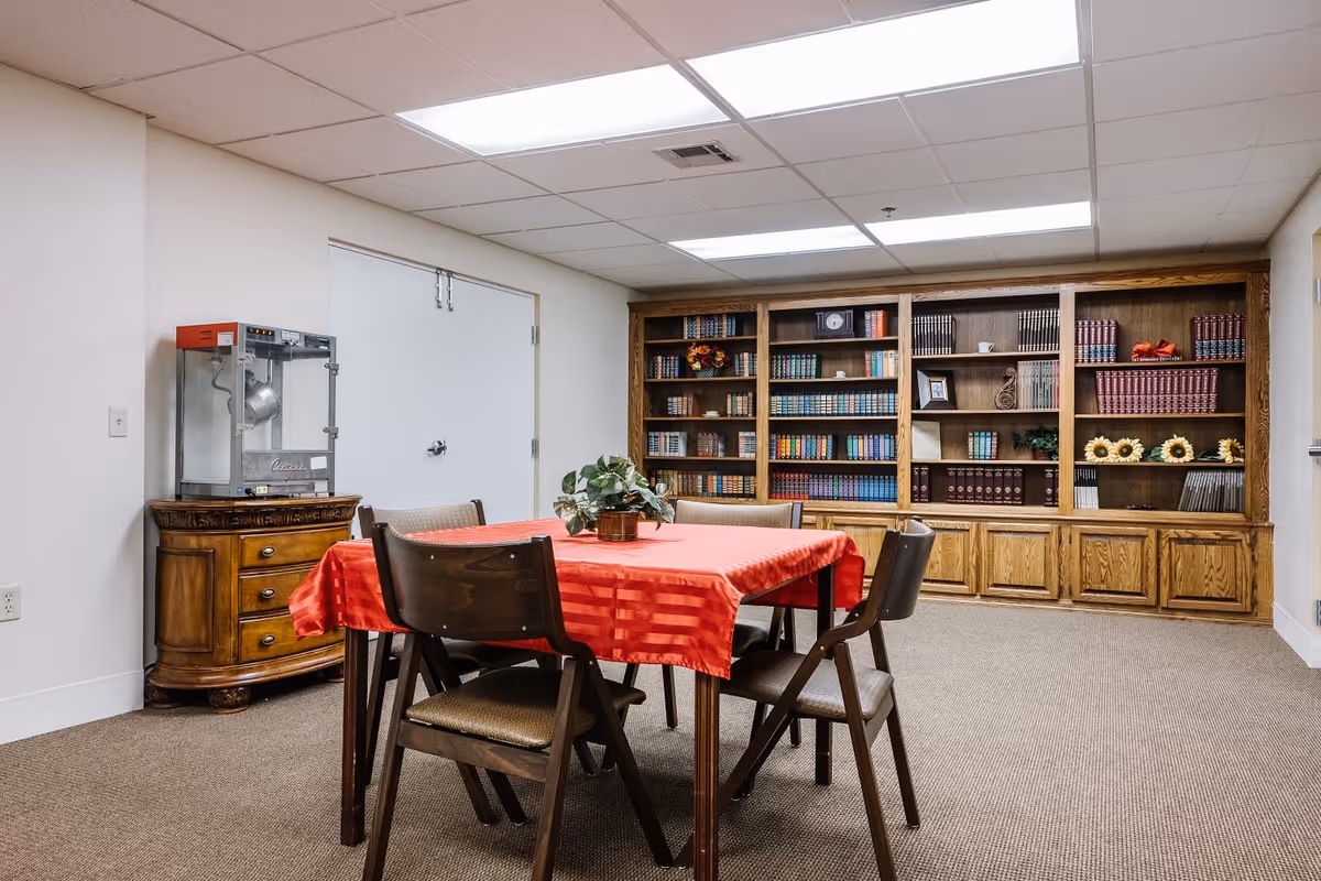 A room with a wooden table covered by a red tablecloth surrounded by four chairs. In the background, there is a large wooden bookshelf filled with books and decorative items. To the left, there is a wooden cabinet with a popcorn machine on top. The room has a carpeted floor and a drop ceiling with fluorescent lights.