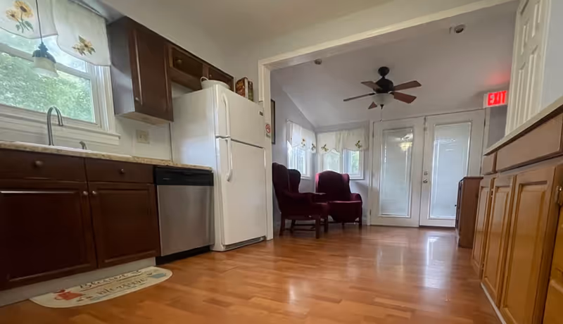 A kitchen with wooden cabinets, a white refrigerator and dishwasher opening into a small sitting area with two red armchairs and double doors.