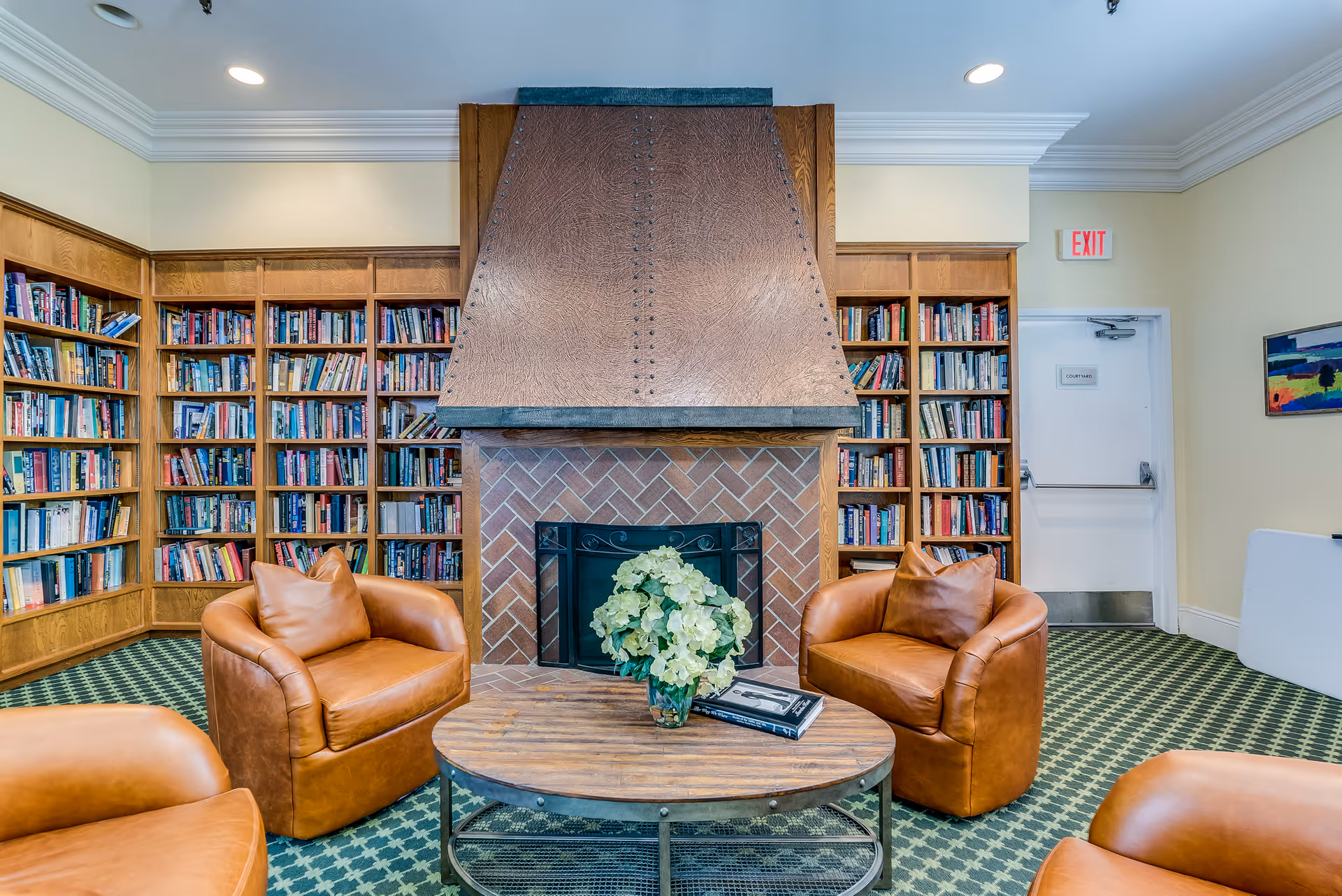 A cozy senior living facility lounge area with four brown leather armchairs arranged around a round wooden coffee table with a vase of flowers and a book. Behind the seating is a large brick fireplace with a copper hood, flanked by built-in wooden bookshelves filled with books. The room has patterned green carpet, cream-colored walls, crown molding, and a white exit door labeled 'Courtyard'.