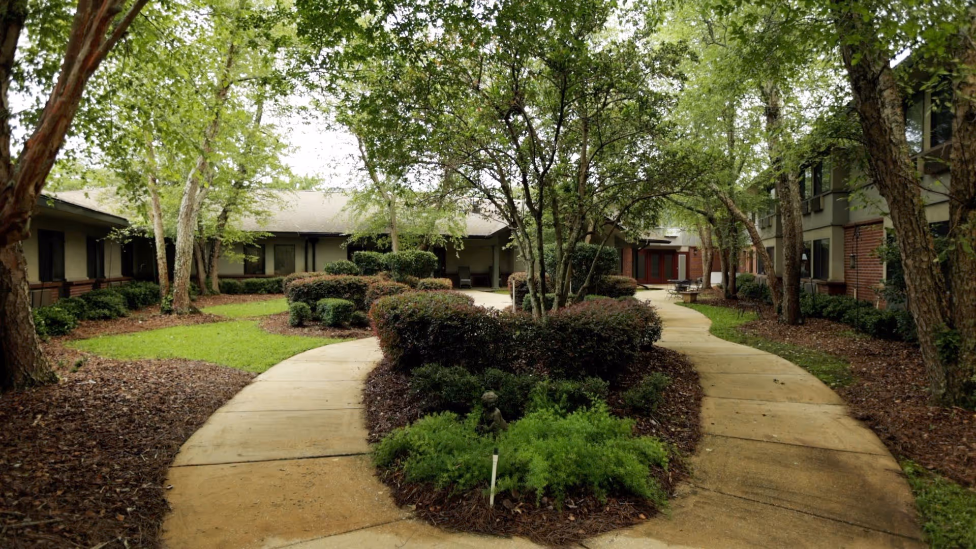 Landscaped courtyard with curved concrete walkways, trimmed bushes and trees between surrounding assisted-living buildings.