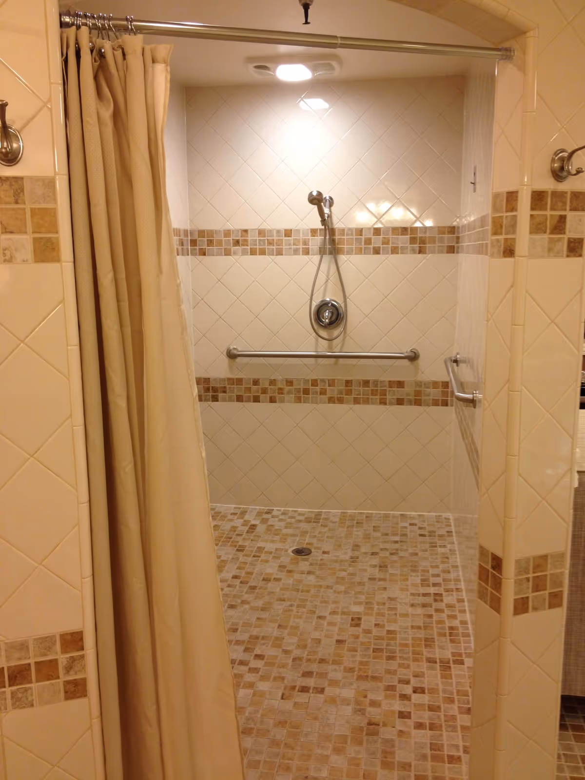 A tiled walk-in shower with beige and brown mosaic floor tiles and matching wall tile accents. The shower has a silver handheld showerhead, a grab bar on the back wall, and a beige shower curtain partially drawn to the side.