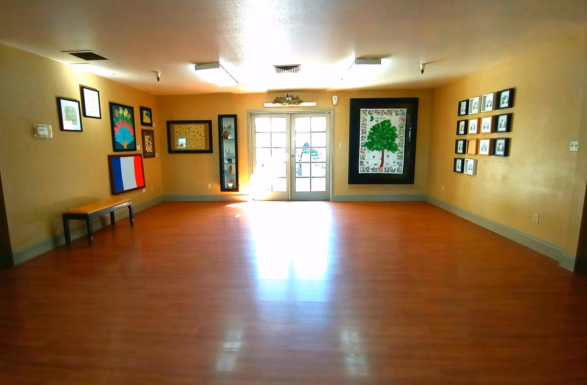 A spacious room with wooden flooring and beige walls decorated with various framed artworks and photographs. There is a bench along the left wall and double glass doors at the far end letting in natural light.