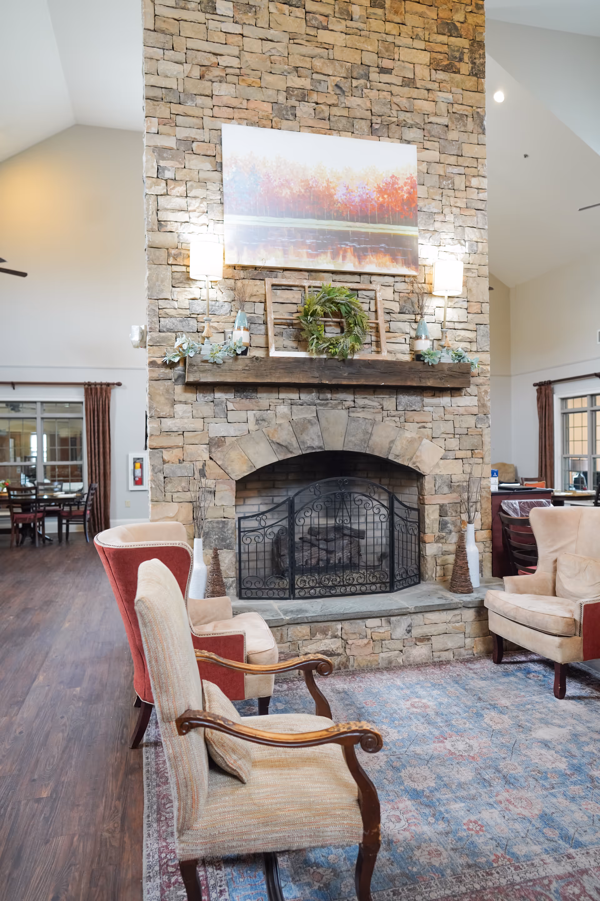 A cozy communal living room featuring a tall stone fireplace with a decorative mantel, armchairs, and a patterned rug.