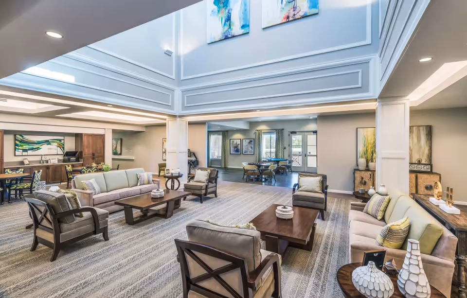 Spacious, well-lit senior living common lounge with sofas, armchairs, coffee tables and a dining area visible in the background under a high ceiling.