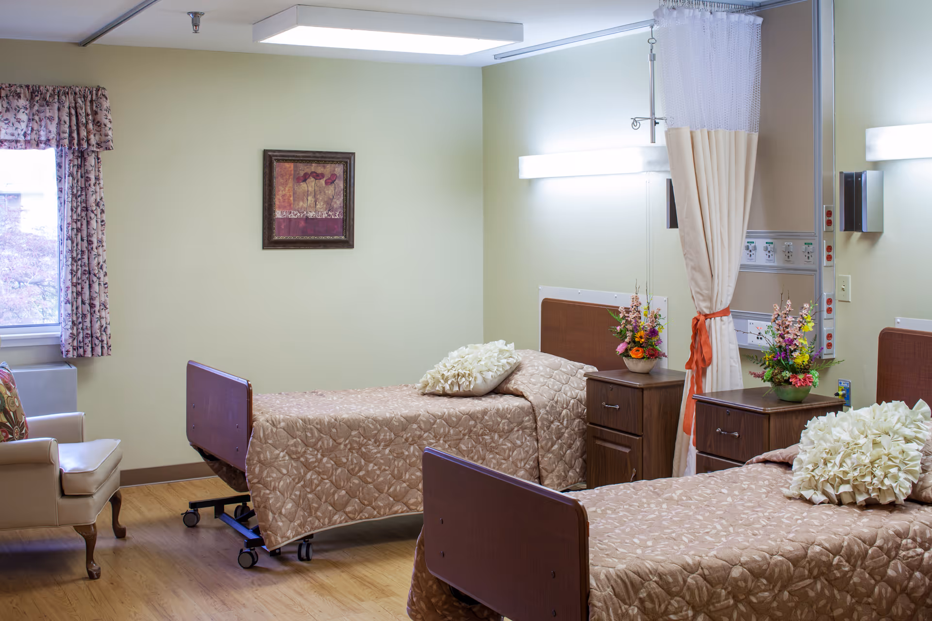 A room with two hospital-style beds covered in beige patterned bedspreads, each with a decorative pillow. Between the beds are two wooden nightstands with flower arrangements. A privacy curtain is partially drawn between the beds. The room has light green walls, a window with floral curtains, a framed picture on the wall, and a beige armchair near the window.