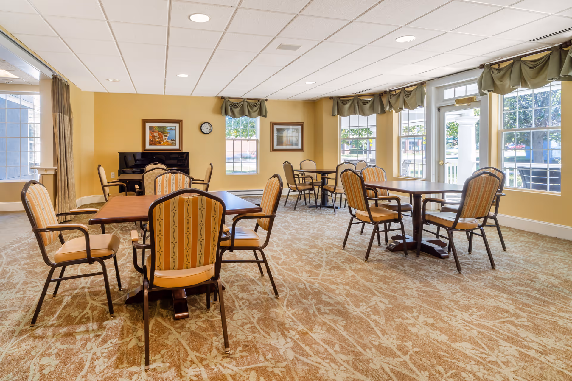 A bright and spacious common dining area with several wooden tables and cushioned chairs arranged neatly. The room has large windows with green valances allowing natural light to fill the space. There is a piano against the far wall, along with framed artwork and a clock. The carpet has a floral pattern and the walls are painted a warm yellow.