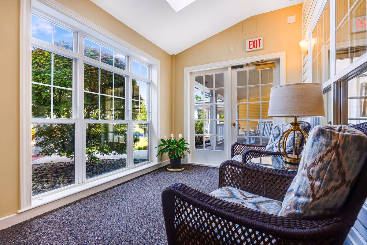 A bright and cozy indoor seating area with two cushioned wicker chairs and a small glass-top table with a lamp. Large windows on the left side let in natural light and offer a view of green trees outside. A potted plant is placed near a glass door with an exit sign above it.