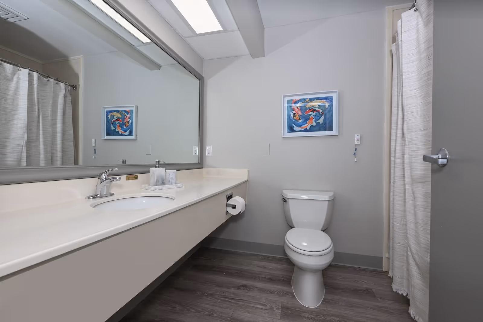 A clean and modern bathroom with a white toilet, a long countertop with an integrated sink, a large mirror above the sink, a shower with a white curtain, and a framed picture of colorful koi fish on the wall.