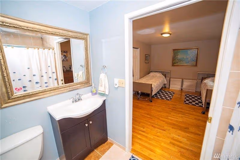 Bathroom vanity and sink with a mirror, looking through a doorway into a bedroom with two single beds on a hardwood floor.