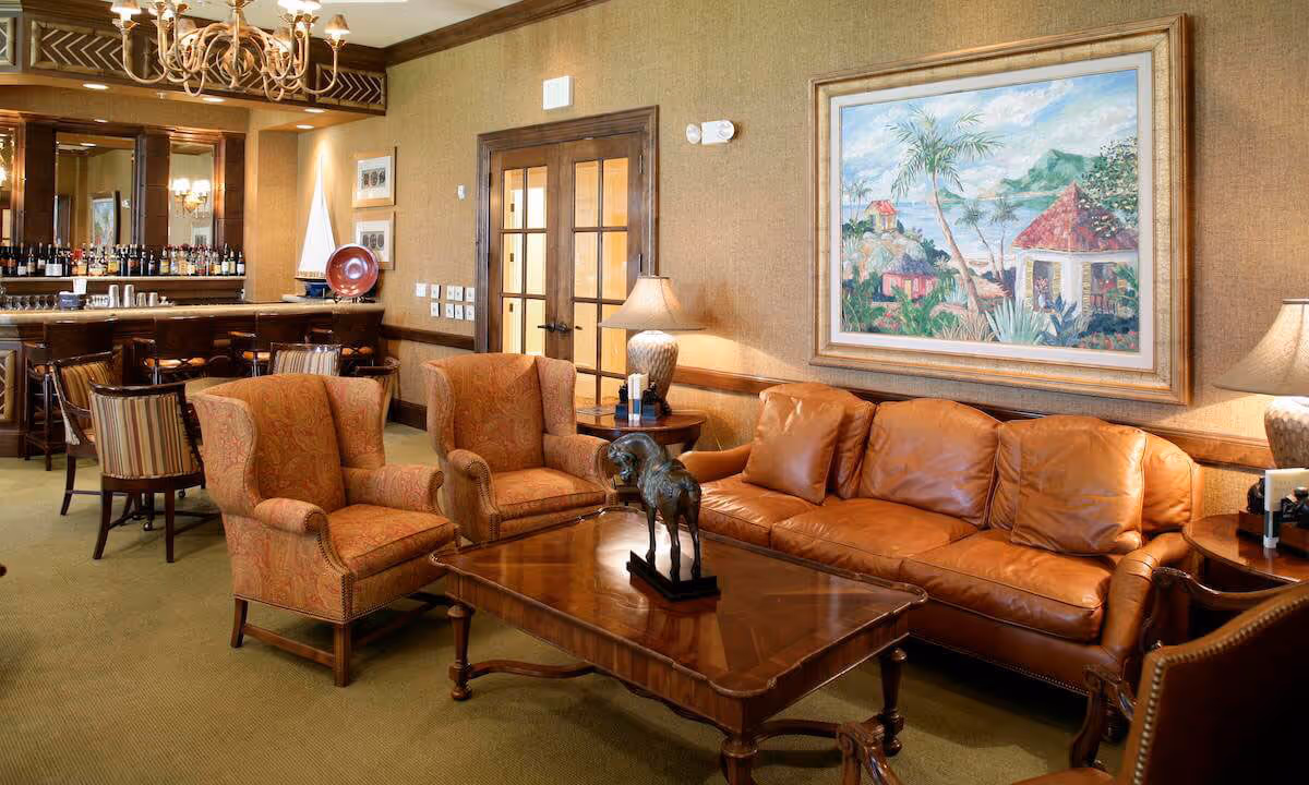 A cozy living room area with a brown leather sofa, two patterned armchairs, a wooden coffee table with a decorative sculpture, and two side tables with lamps. In the background, there is a bar area with high chairs and a chandelier hanging from the ceiling. A large framed painting depicting a tropical scene hangs on the wall above the sofa.