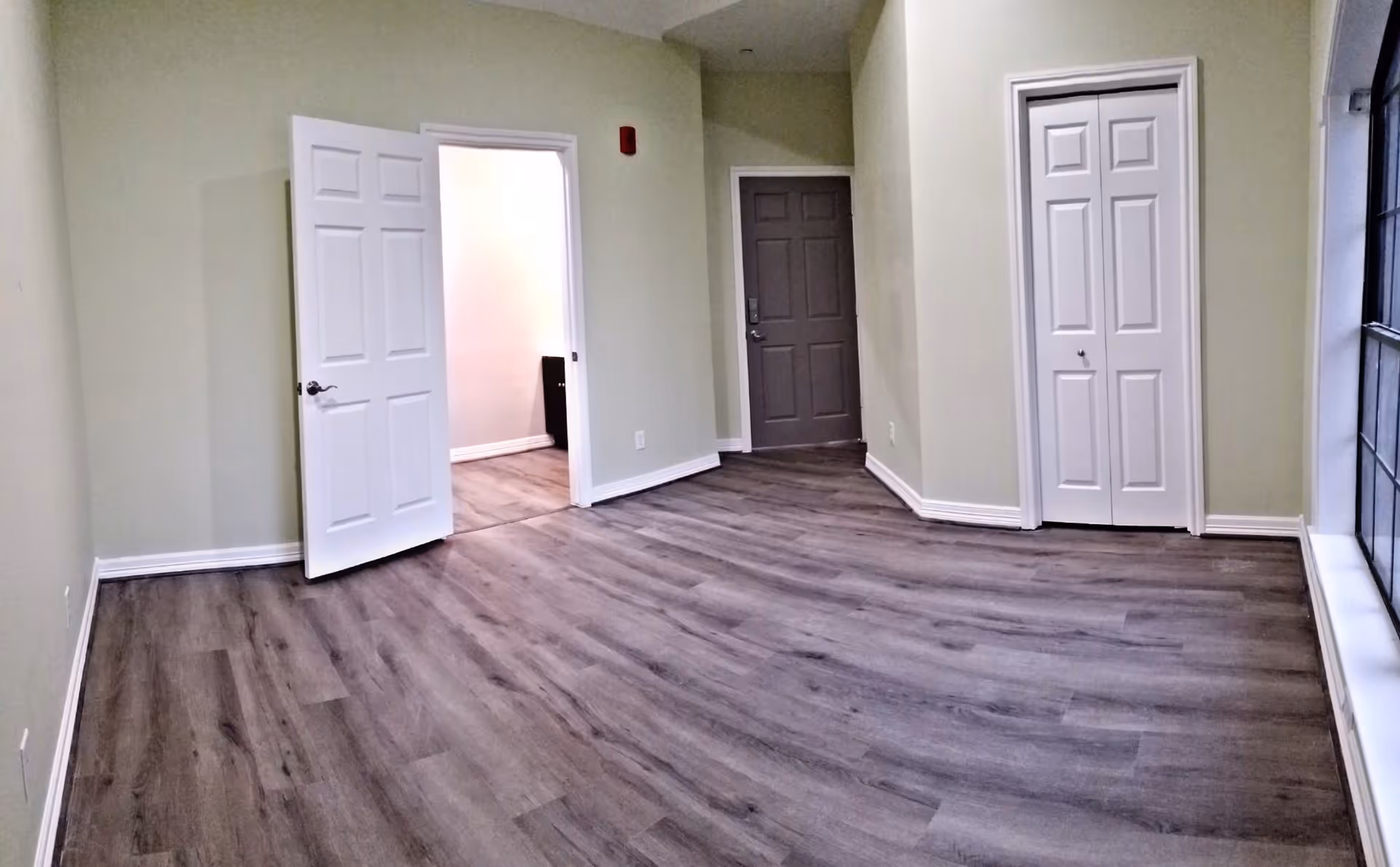Empty room with light green walls and wood flooring, featuring a white door open to another room, a closed white closet door, a gray entrance door, and a large window on the right side.