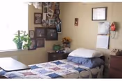 Small bedroom with a single bed covered by a patchwork quilt, a nightstand, framed photos on the wall, and a window with a potted plant.
