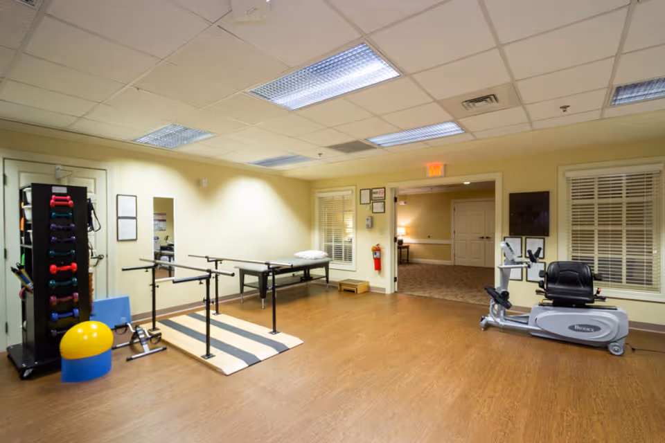 A physical therapy or rehabilitation room with exercise equipment including a rack of colorful dumbbells, parallel bars for walking exercises, a padded therapy table with a pillow, a yellow and blue exercise ball, and a stationary recumbent exercise bike. The room has wooden flooring, beige walls, fluorescent ceiling lights, and a doorway leading to another carpeted room.