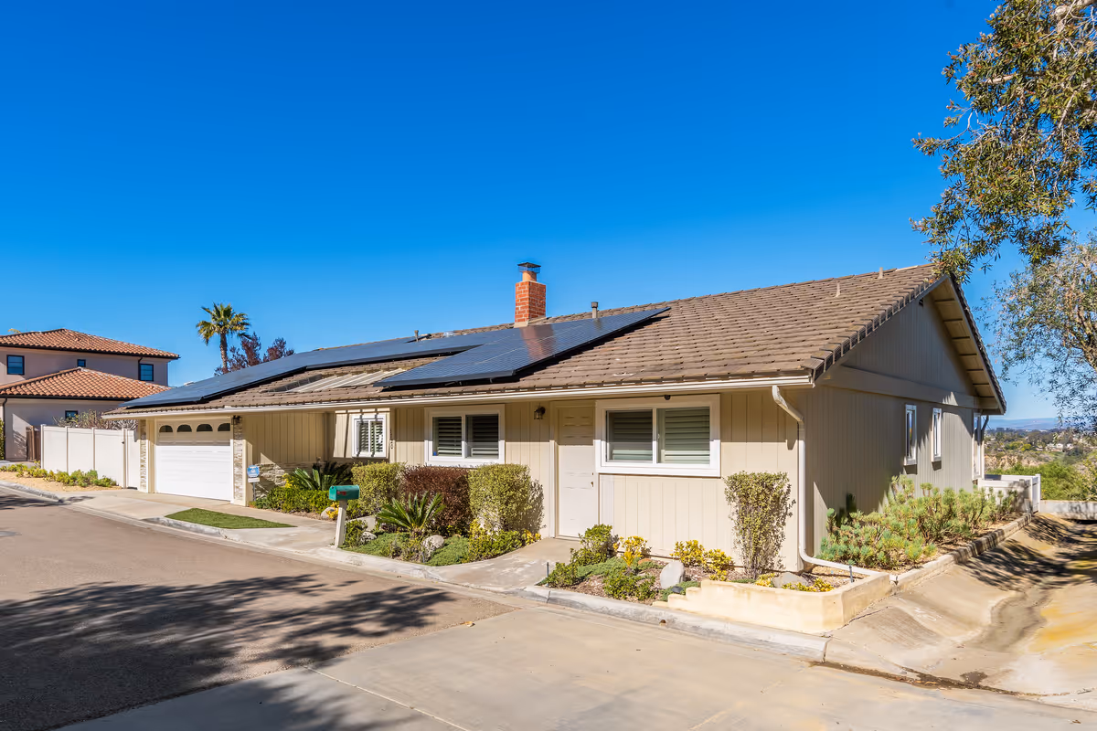 Single-story beige house with solar panels on the roof, an attached two-car garage, and a small landscaped front yard under a clear blue sky.