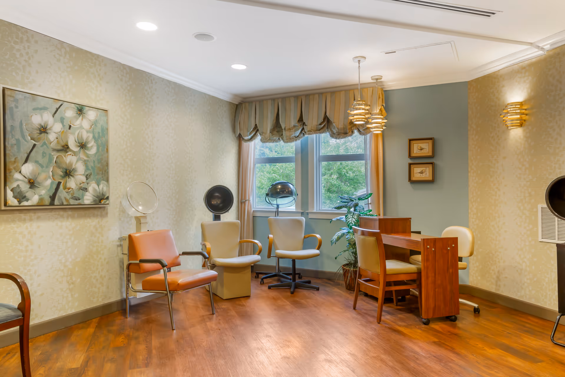 Interior room with wooden flooring and light-colored walls featuring floral artwork. The room contains several chairs, including salon-style chairs with hair dryers, a small wooden table with chairs, and two windows with striped valances showing greenery outside.