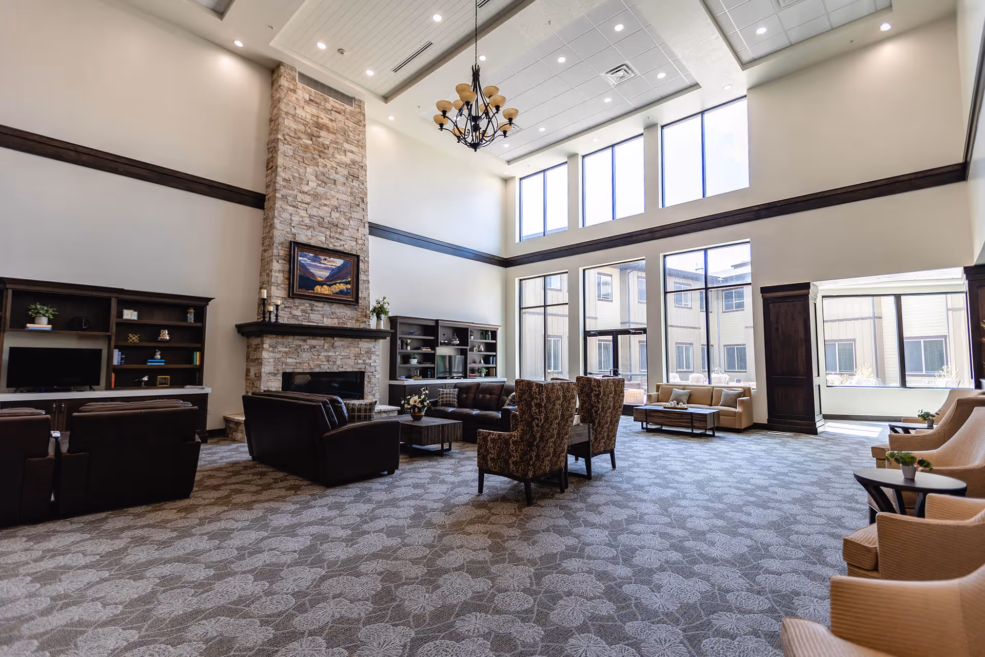 Spacious senior living facility common area with high ceilings, large windows allowing natural light, a stone fireplace with a painting above it, multiple seating arrangements including leather sofas, patterned armchairs, and beige chairs with small tables, and built-in shelves with decorative items and a television.