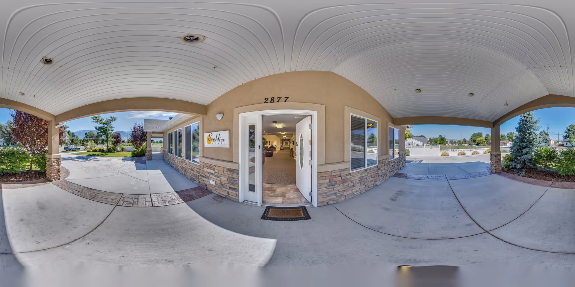 Covered entrance area of BeeHive Homes of Provo with a beige exterior wall, stone accents, and a white door open to the interior. The address number 2877 is above the door, and there is a sign with the BeeHive Homes logo on the wall. The area is surrounded by landscaped greenery and trees under a clear blue sky.