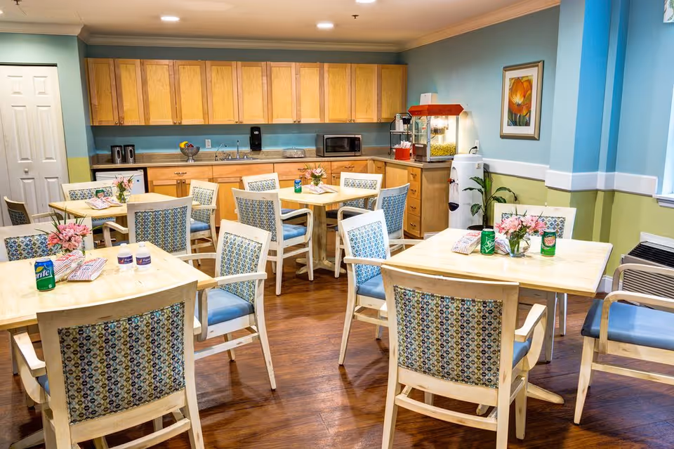 Bright communal dining room with wooden tables and patterned chairs arranged near a kitchenette and popcorn machine.