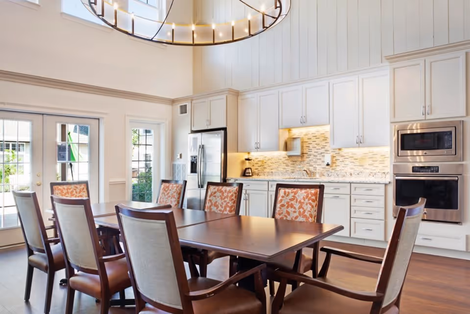 A bright and spacious kitchen and dining area with a large wooden dining table surrounded by eight chairs, some with floral patterned upholstery. The kitchen features white cabinetry, a stainless steel refrigerator, built-in microwave and oven, and a tiled backsplash. Large windows and glass doors allow natural light to fill the room.