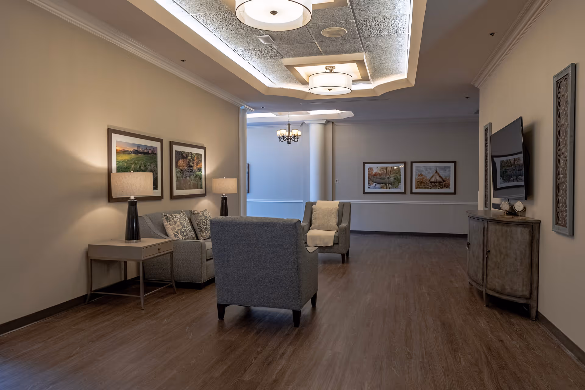 Well-lit nursing home lounge with upholstered chairs and sofa, side tables with lamps, framed artwork, and a TV on a console.