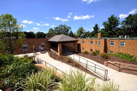 Outdoor view of a senior living facility with a brick building, a covered entrance, a wheelchair ramp, and greenery including bushes and trees under a blue sky with scattered clouds. Two people are visible near the entrance, one pushing the other in a wheelchair.