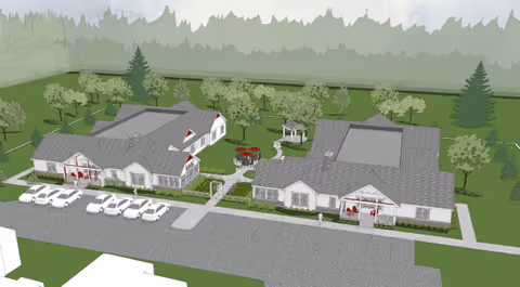 Aerial view of a senior living facility named Willow Creek West AFC showing multiple connected buildings with gray roofs, surrounded by green lawns, trees, and pathways. There are several white cars parked in front of the buildings, and outdoor seating areas with red umbrellas are visible. The background features a forested area.