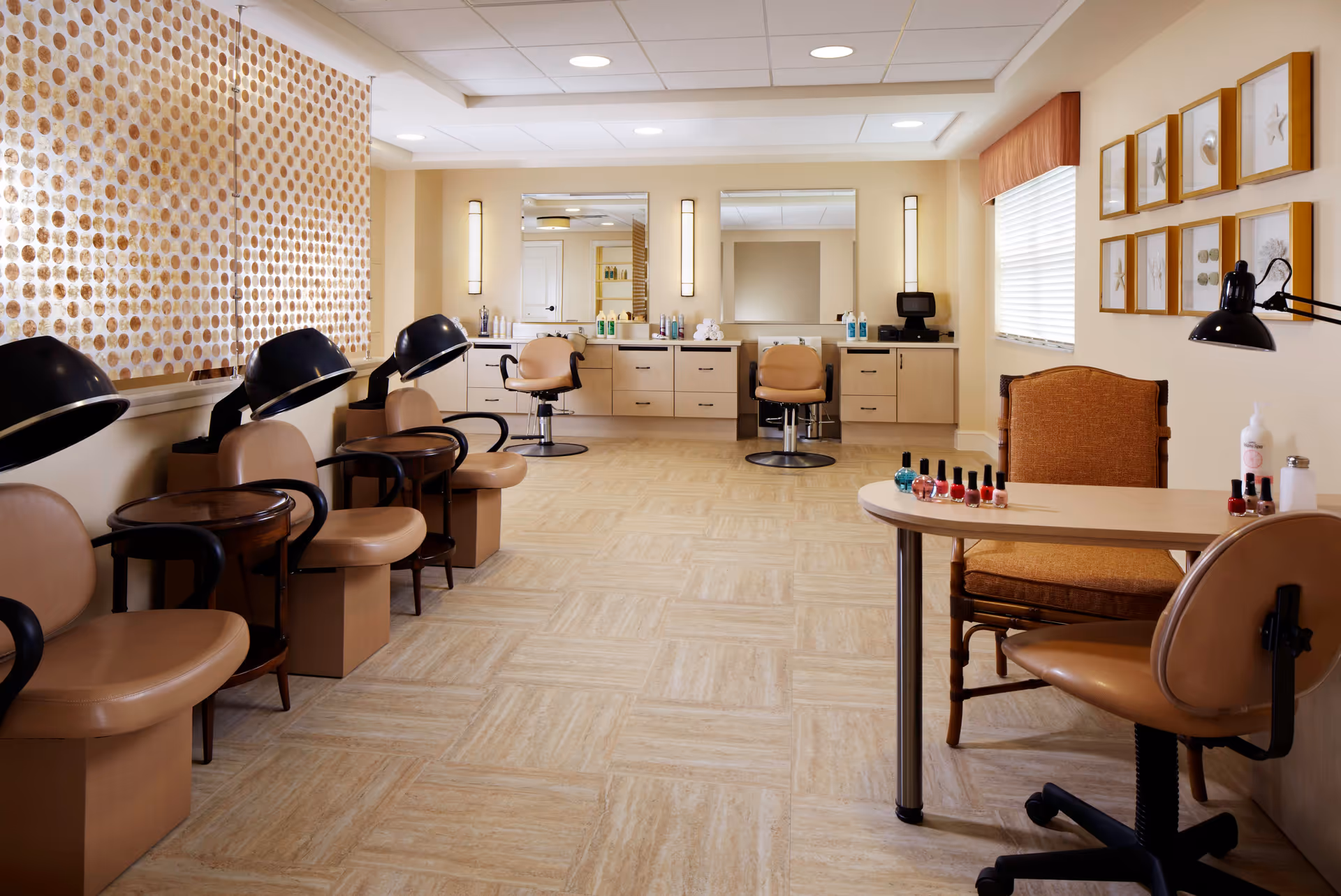 A well-lit salon area in a senior living facility with multiple beige salon chairs, hair dryers, and a manicure table with nail polish bottles. The room has light-colored walls, framed artwork, and large mirrors above a counter with various hair care products.