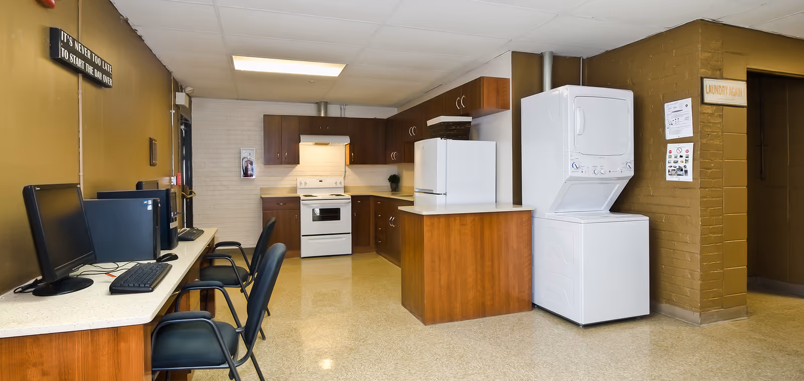 A communal room featuring a kitchen area with wooden cabinets, a white stove, microwave, and refrigerator. To the right, there is a stacked washer and dryer unit. On the left side, there is a counter with two desktop computers and chairs. The walls are painted brown and white, with a sign that reads 'IT'S NEVER TOO LATE TO START THE DAY OVER' and another sign near the laundry area that says 'LAUNDRY AGAIN'.