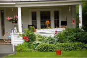 Front porch of a house with potted and hanging plants, a bicycle, and French doors leading inside.