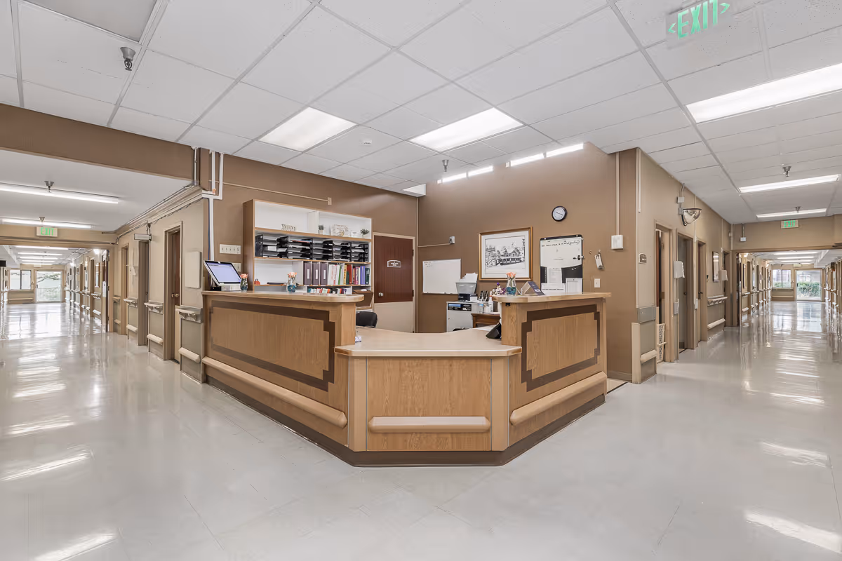 A central nurses' station and reception desk in a long, well-lit senior care facility corridor.