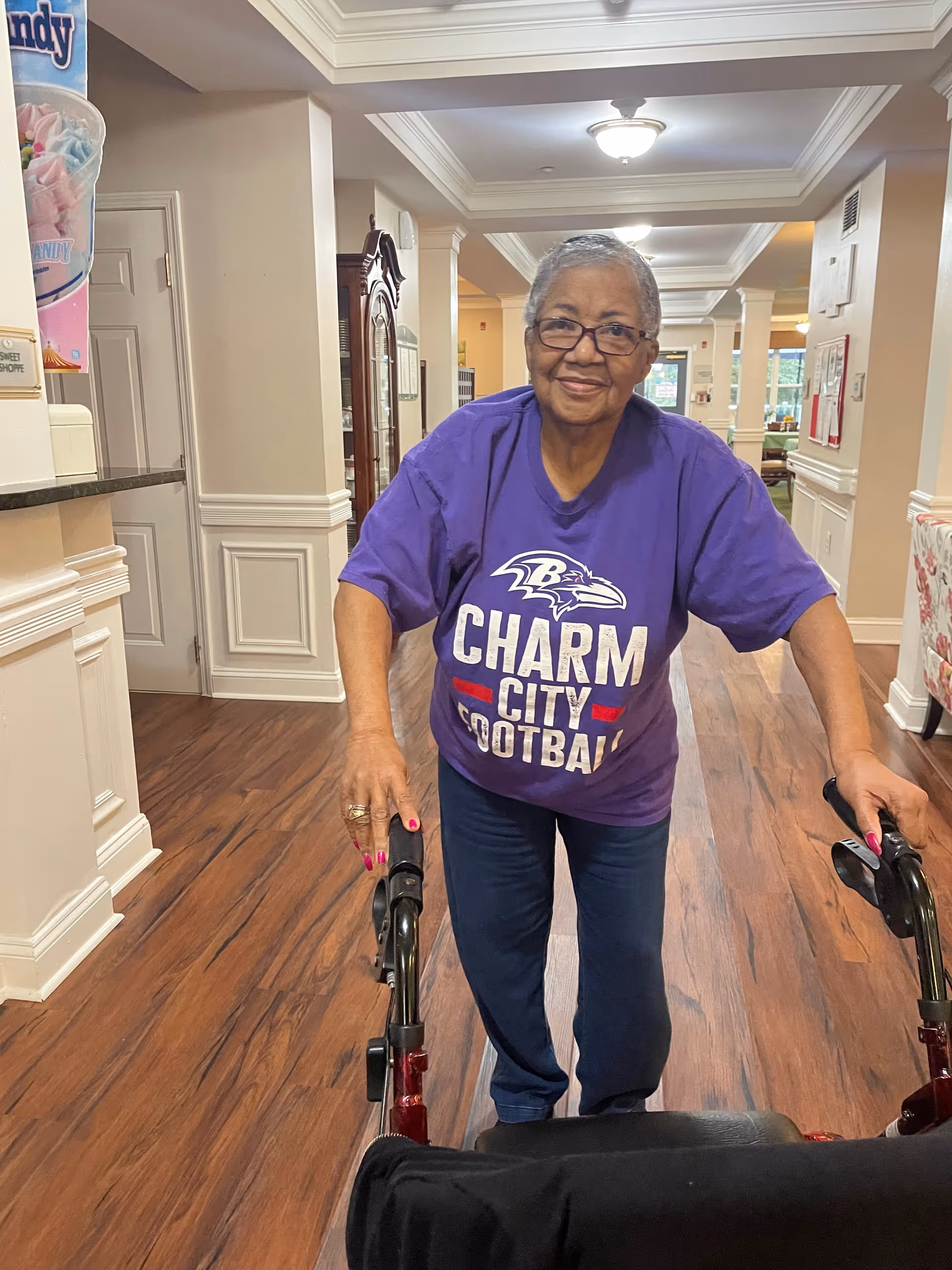 An elderly woman wearing glasses and a purple Baltimore Ravens Charm City Football t-shirt is standing indoors using a walker. The setting appears to be a well-lit hallway with wood flooring and white trim, likely inside an assisted living facility.