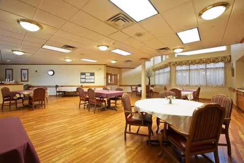 A spacious dining room with several round and rectangular tables covered with tablecloths, surrounded by wooden chairs with cushioned seats and backs. The room has wooden flooring, large windows with curtains, and ceiling lights providing bright illumination. There are decorative elements on the walls and a bulletin board near the back door.