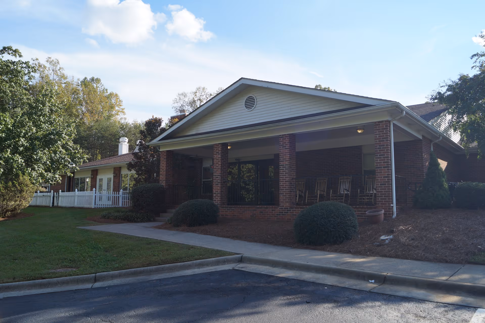 Exterior view of a single-story brick building with a covered porch featuring several rocking chairs. The building is surrounded by greenery including bushes and trees, with a paved driveway and sidewalk in front.