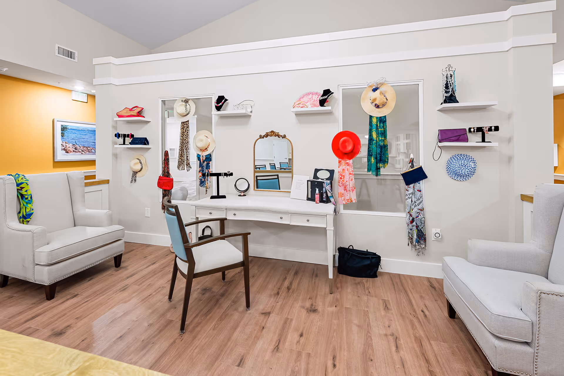 A bright and inviting interior room with a white vanity table and mirror, surrounded by shelves and hooks displaying various hats, scarves, and handbags. Two comfortable armchairs are positioned on either side of the vanity, and the room features light wood flooring and white walls with a yellow accent wall in the background.