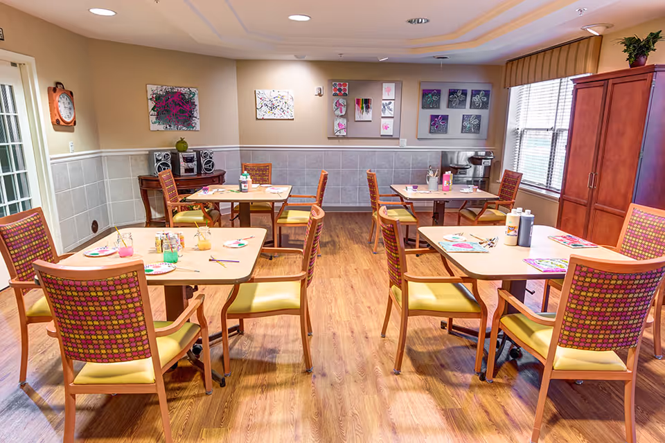 Bright dining/activity room with several tables and chairs arranged for arts-and-crafts activities.