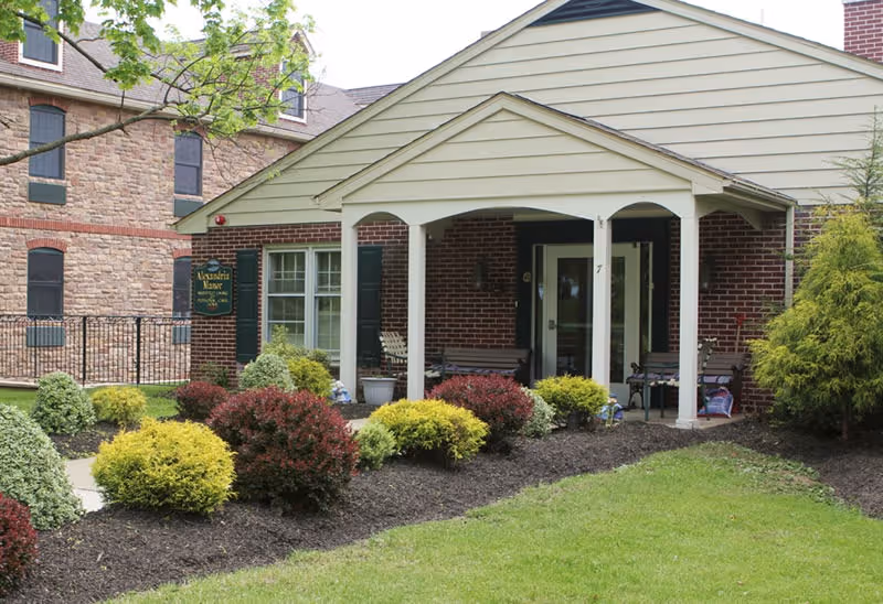 Front entrance of Alexandria Manor, a brick building with a covered porch supported by white columns. The porch has benches and potted plants. In front of the building is a landscaped garden with various shrubs and green grass.