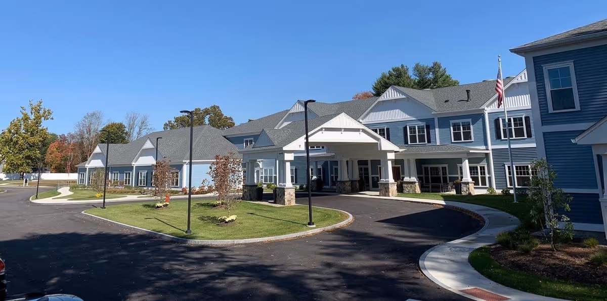 Exterior view of The Branches of Framingham senior living facility showing a large, two-story building with blue siding and white trim. The building features a covered entrance with stone pillars, a curved driveway, street lamps, and landscaped greenery including small trees and bushes. The sky is clear and blue.