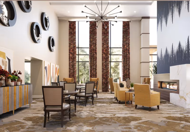 Bright high‑ceiling communal dining and lounge area with round tables, chairs, yellow armchairs, tall windows, and a fireplace.