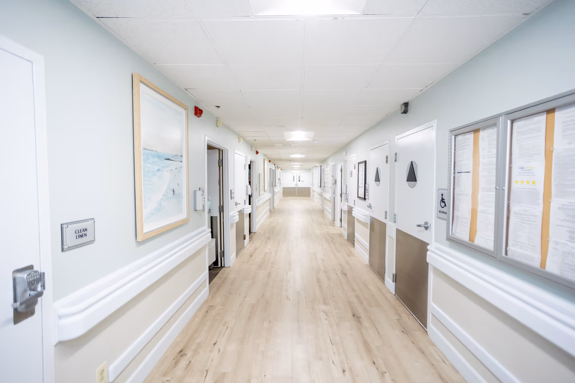 A bright, clean hallway in a senior living facility with light wood flooring, white walls, and multiple doors on both sides. There are handrails along the walls, a framed picture, and a bulletin board with notices. The hallway is well-lit with ceiling lights.