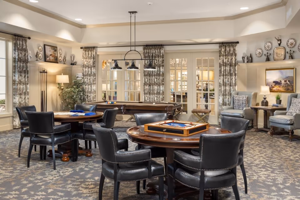 A cozy and well-lit common area in an independent living community featuring round wooden tables with black leather chairs, a pool table in the background, patterned curtains on large windows, decorative plates and artwork on the walls, and comfortable armchairs with side tables and lamps.