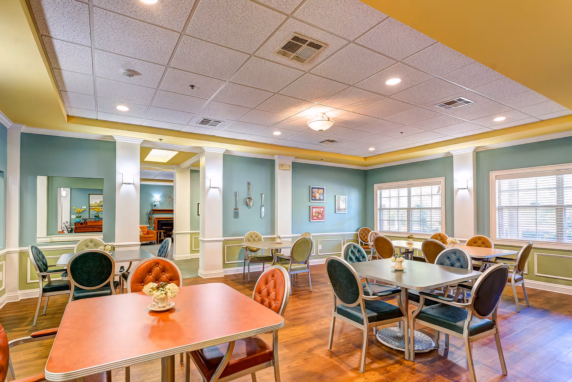 Bright dining room with multiple tables and upholstered chairs, wood floors, and large windows in a senior living facility.